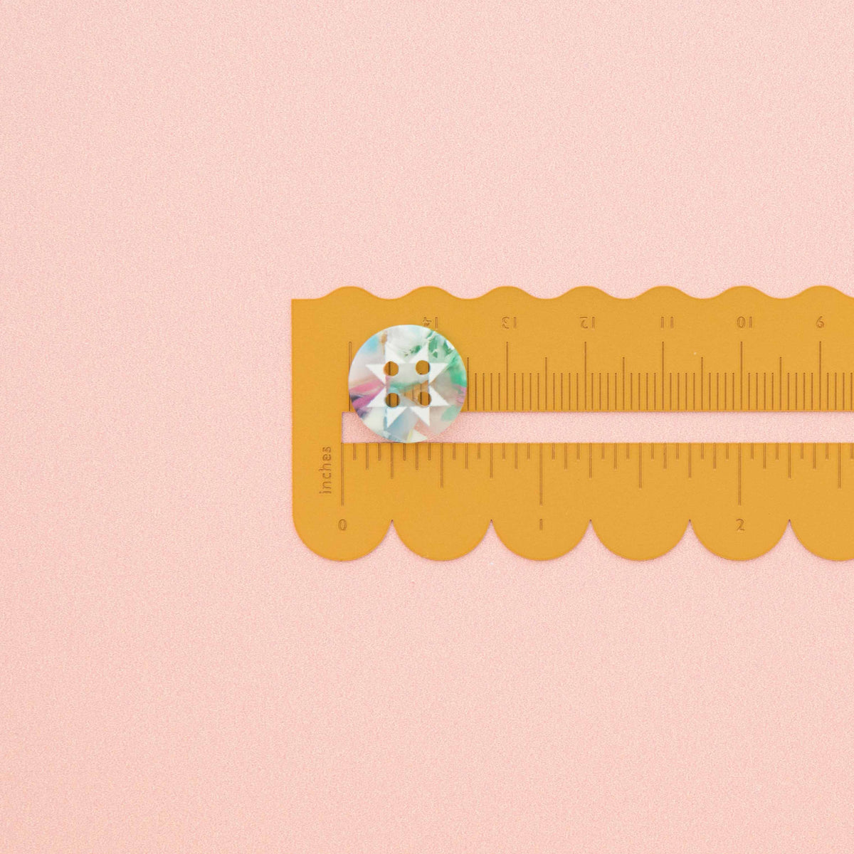 A round four-hole button with a pastel marbled pattern in blue, green, and pink, placed on a yellow scalloped ruler against a solid pink background.