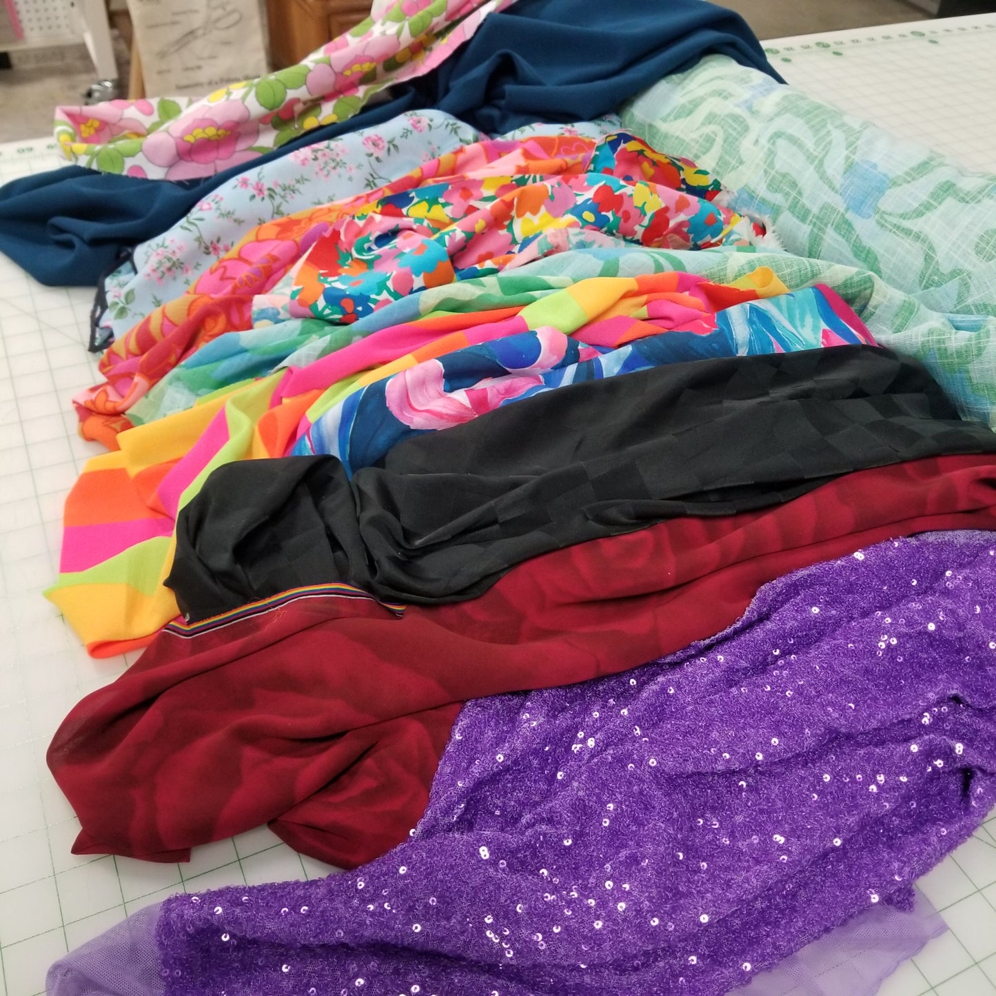 Stack of colorful fabrics including floral, sequined, and solid colors on a white surface. Designer Deadstock