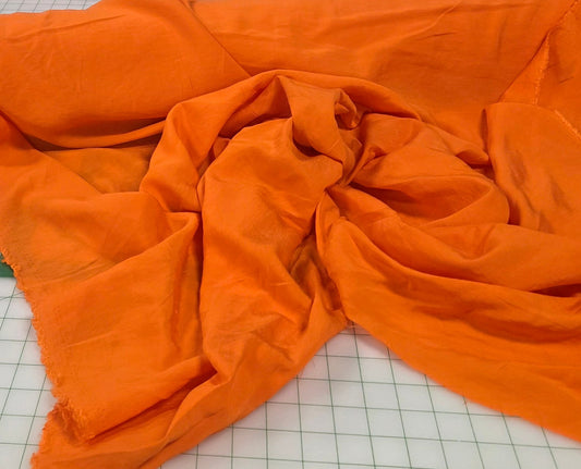 Orange fabric draped over a white surface with measurement markings, surrounded by various items in a store setting. Orange rayon challis textured.