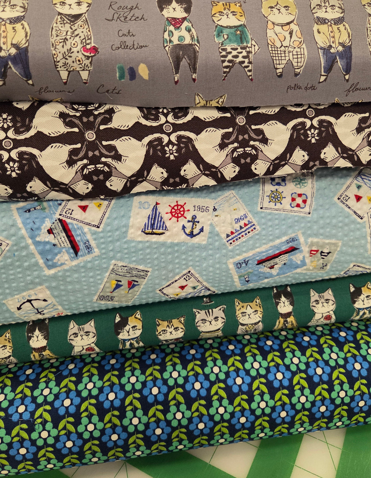A stack of five patterned fabric rolls displayed on a cutting mat. The fabrics feature various designs including cartoon cats in clothing, a black and white pattern, nautical ship and sailboat motifs, and blue floral patterns.