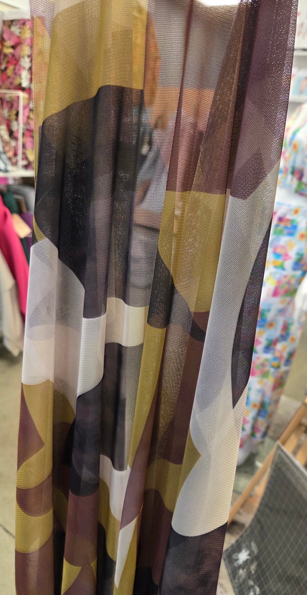 A close-up of a sheer curtain panel featuring an abstract camouflage pattern in brown, beige, white, and black, hanging in a retail store.