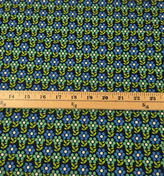 A strip of fabric with a repeating blue, green, and white floral pattern, shown vertically next to a wooden ruler on a green grid cutting mat.