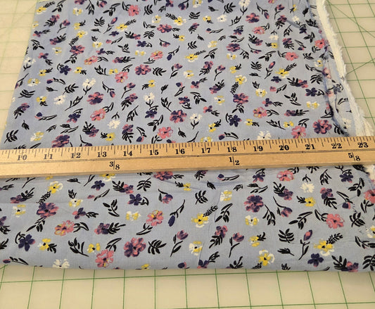 A square piece of light blue floral fabric with a small-scale multicolor print, shown next to a wooden yardstick on a green grid cutting mat.
