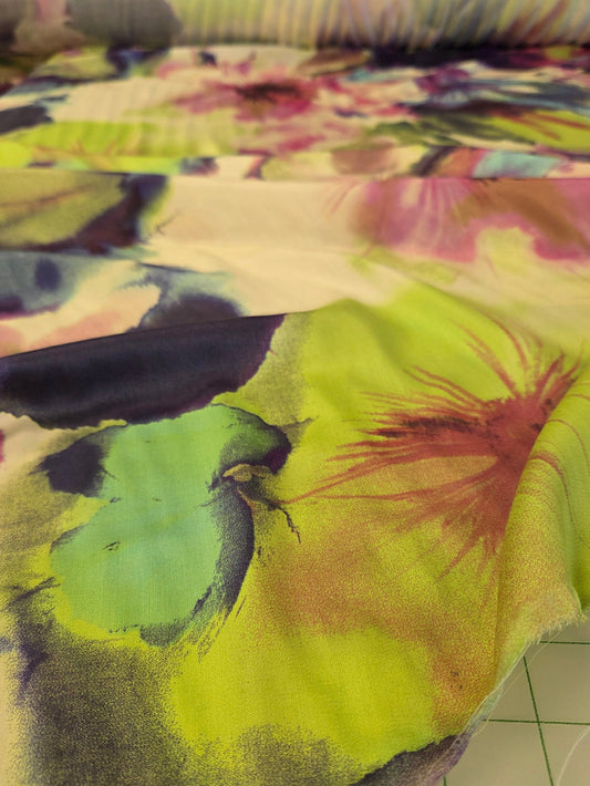 A close-up of a colorful fabric with a watercolor floral pattern in lime green, pink, purple, and blue, draped next to a white cutting mat with grid lines.
