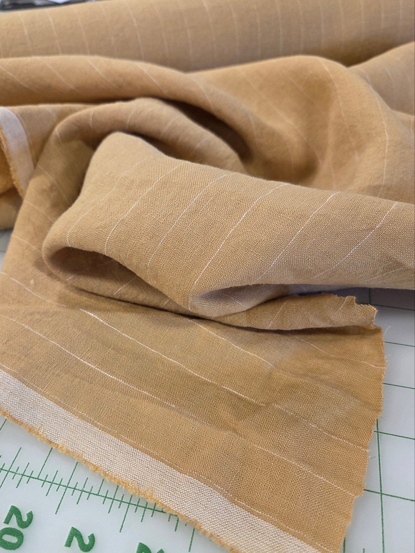 A close-up of a beige fabric with thin white vertical stripes draped over a white gridded cutting mat with green measurement markings.