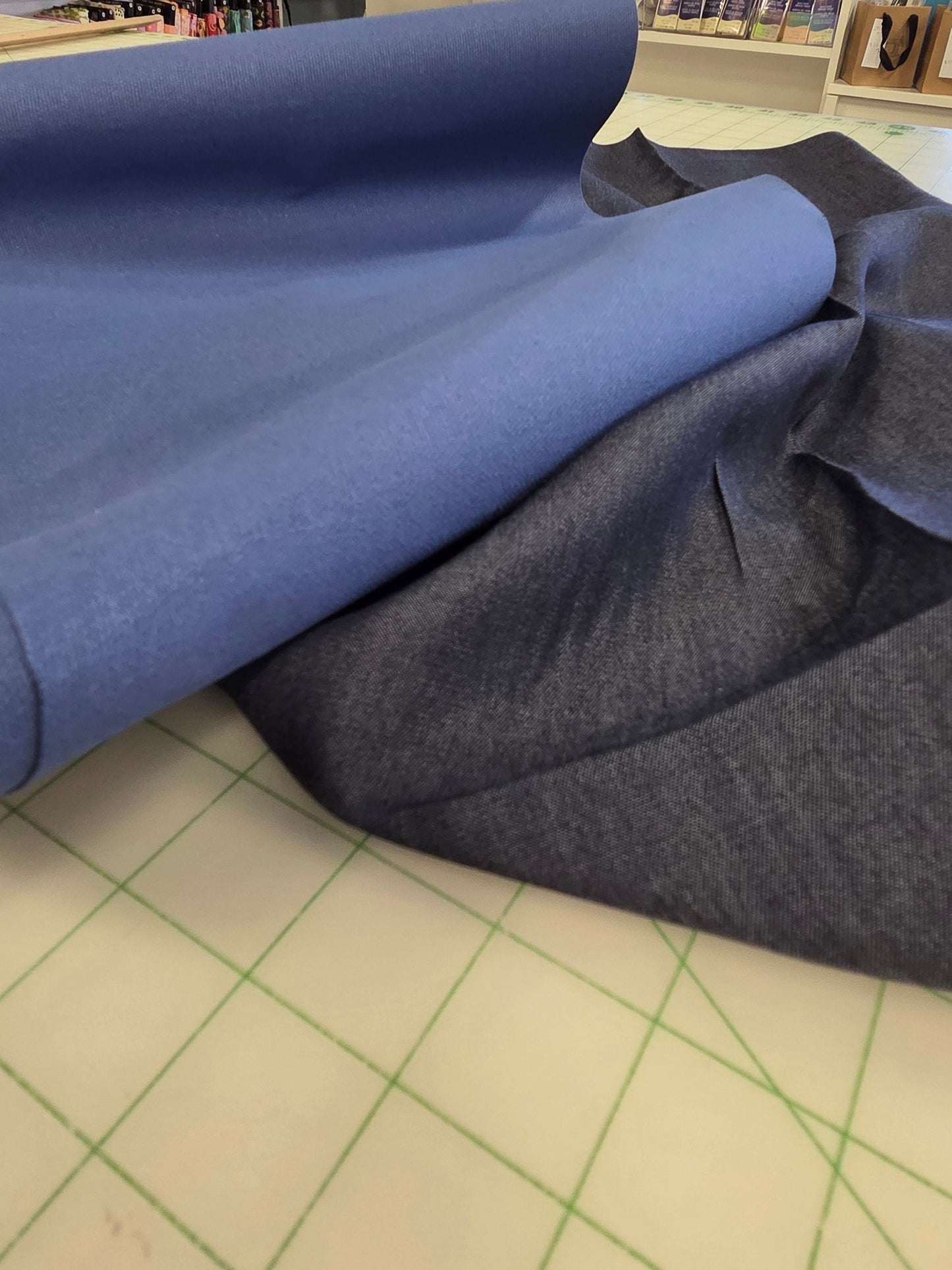A roll of medium blue fabric partially unrolled onto a light-colored cutting mat inside a sewing or fabric store, with shelves and sewing machines visible in the background.