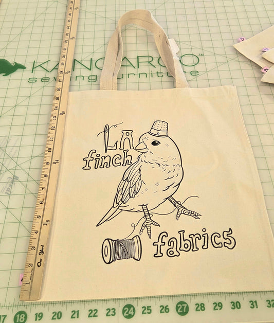 A beige canvas tote bag with a black line art illustration of a finch and the text 'LA finch fabrics' is displayed on a green cutting mat on a worktable. A wooden ruler is positioned next to the bag.