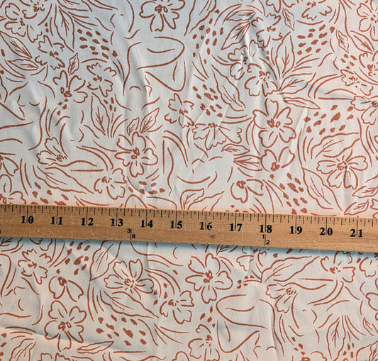 A close-up of a white fabric with a dusty rose or terracotta floral and leaf pattern, shown with a wooden ruler across the middle for scale.