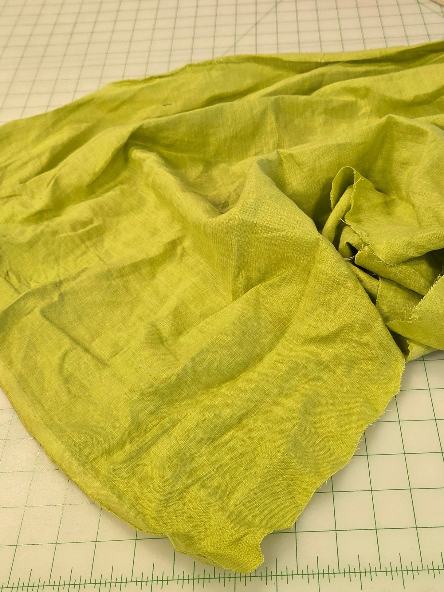A lime green linen fabric scarf or shawl is laid out on a white gridded cutting mat on a sewing table, surrounded by sewing tools and other fabrics.
