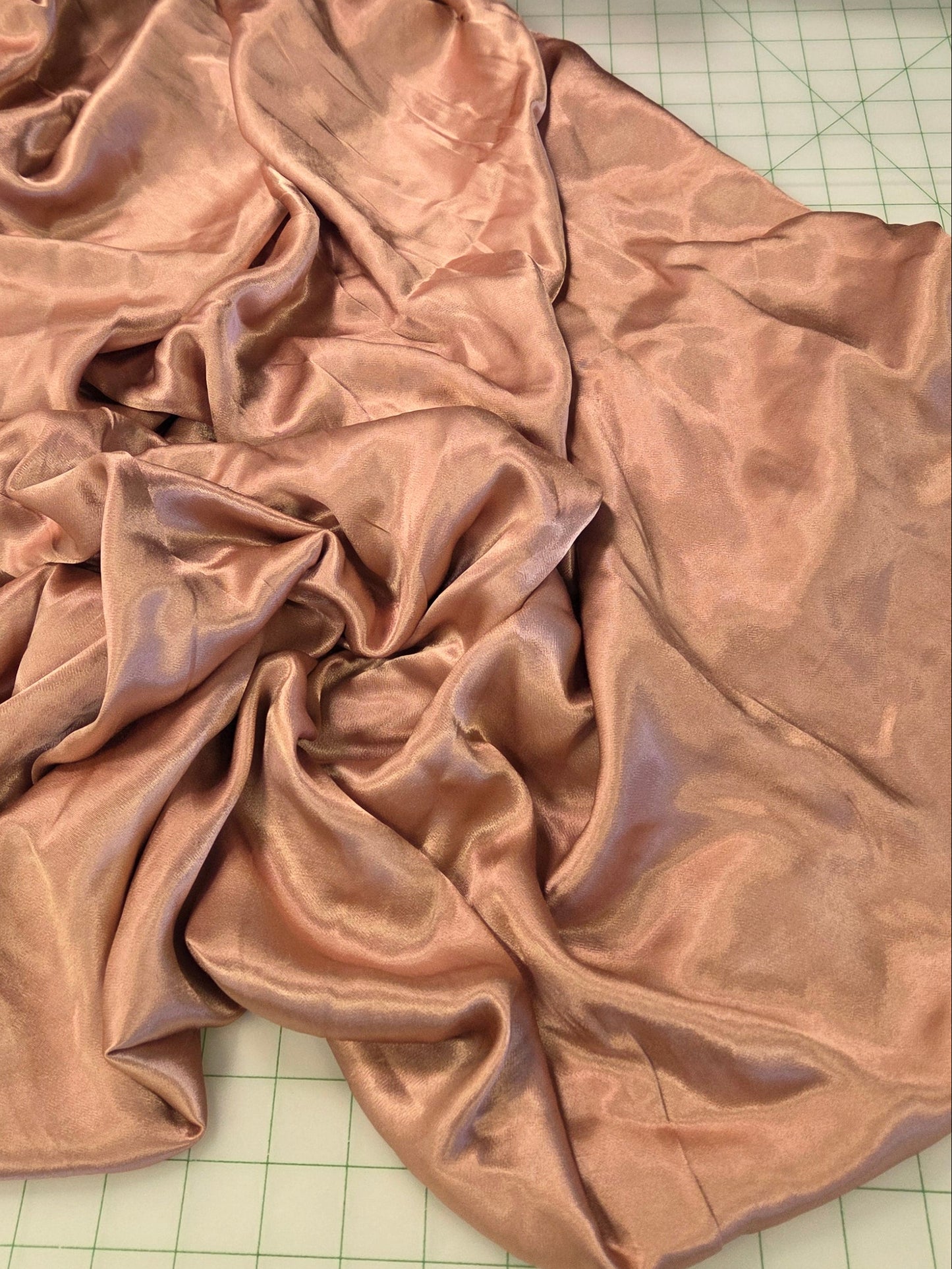 A piece of shiny, draped satin fabric in a dusty rose or bronze color spread out on a green grid cutting mat, with a roll of black fabric in the background. satin shiny cupor pink