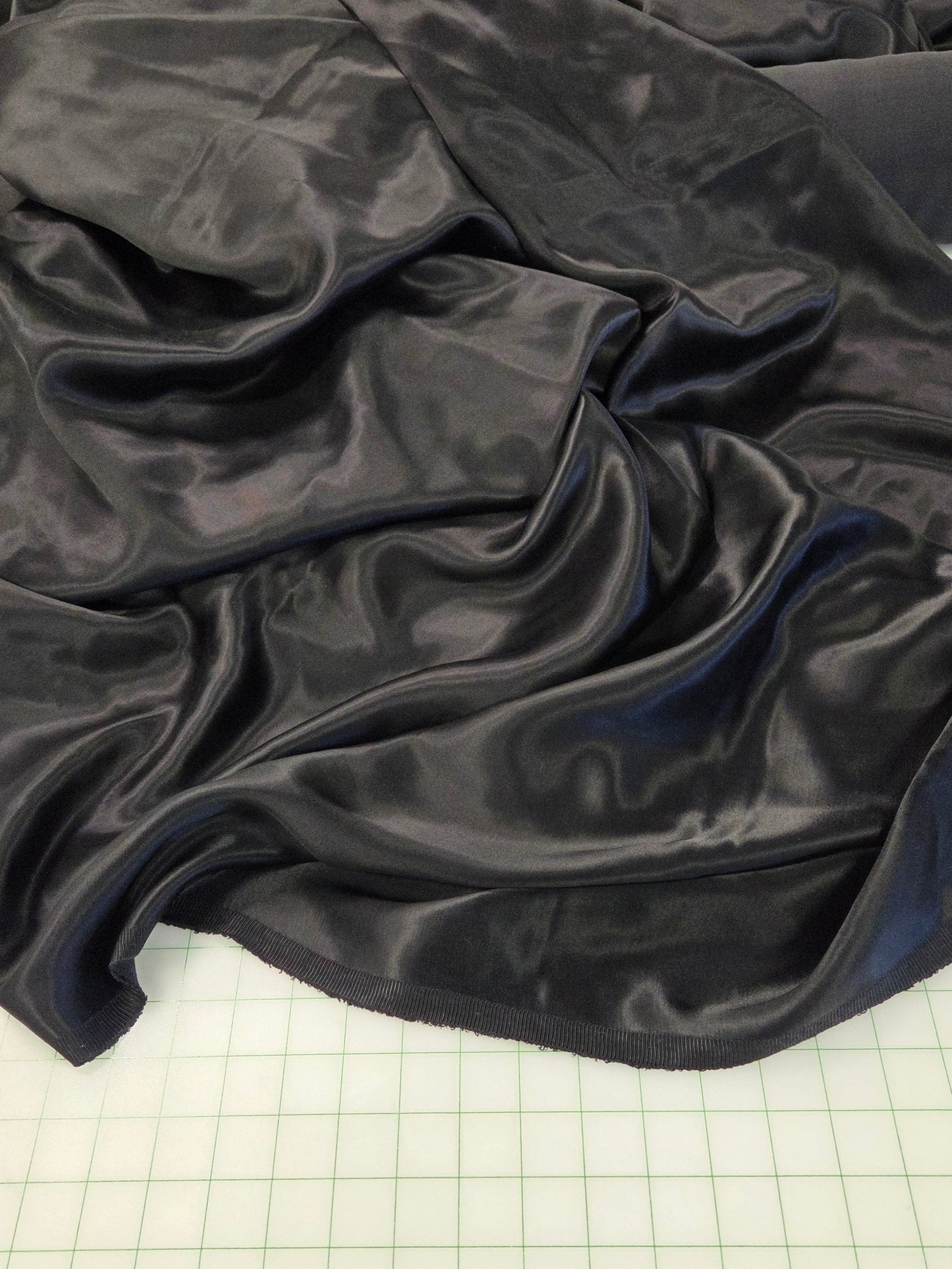 A draped piece of shiny black satin or silk fabric spread across a green gridded cutting mat, with a roll of matching black fabric visible in the background.