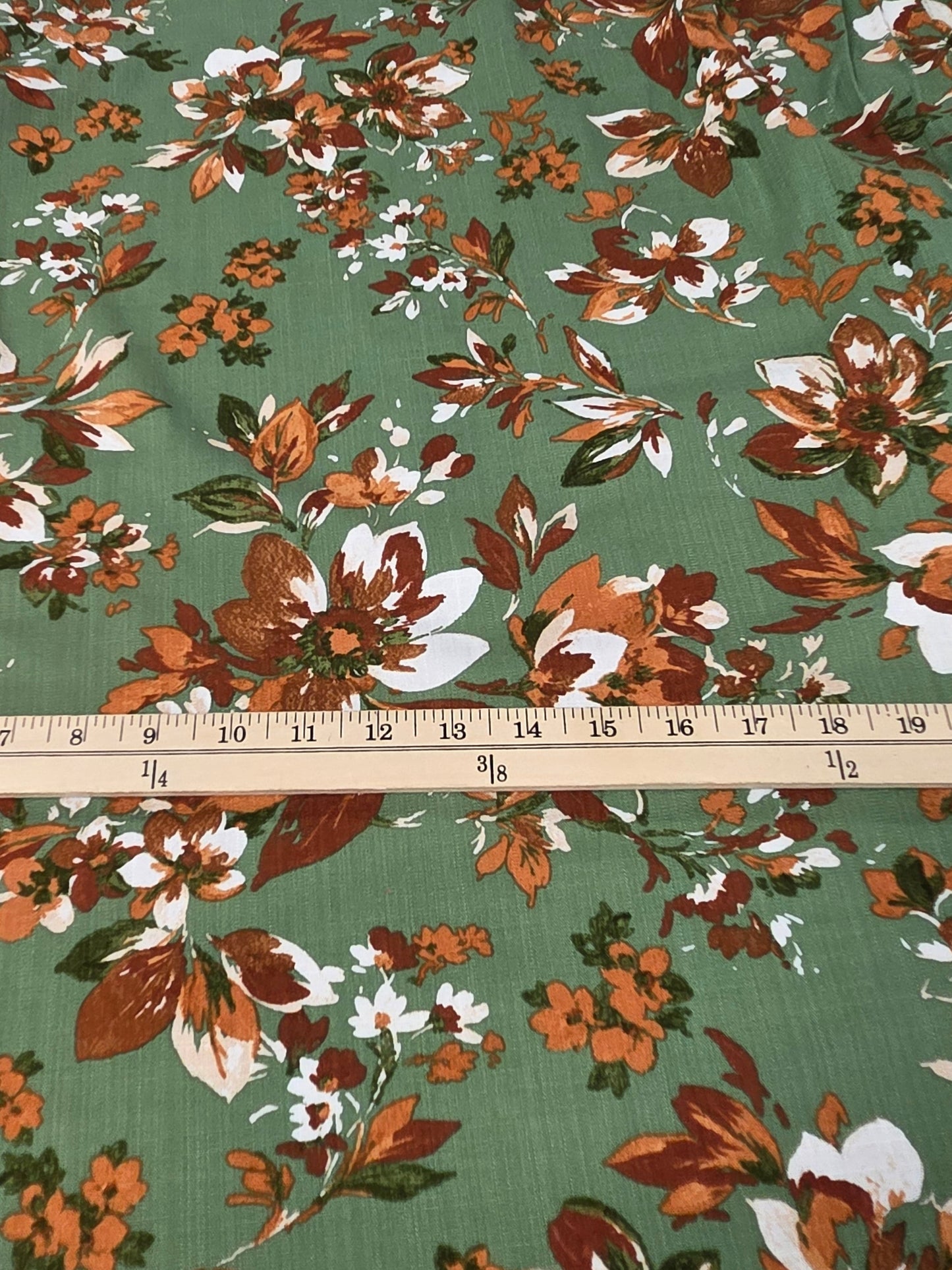 A piece of green fabric with a vintage floral print in orange, brown, and white, laid flat on a cutting mat with a wooden ruler showing its width.