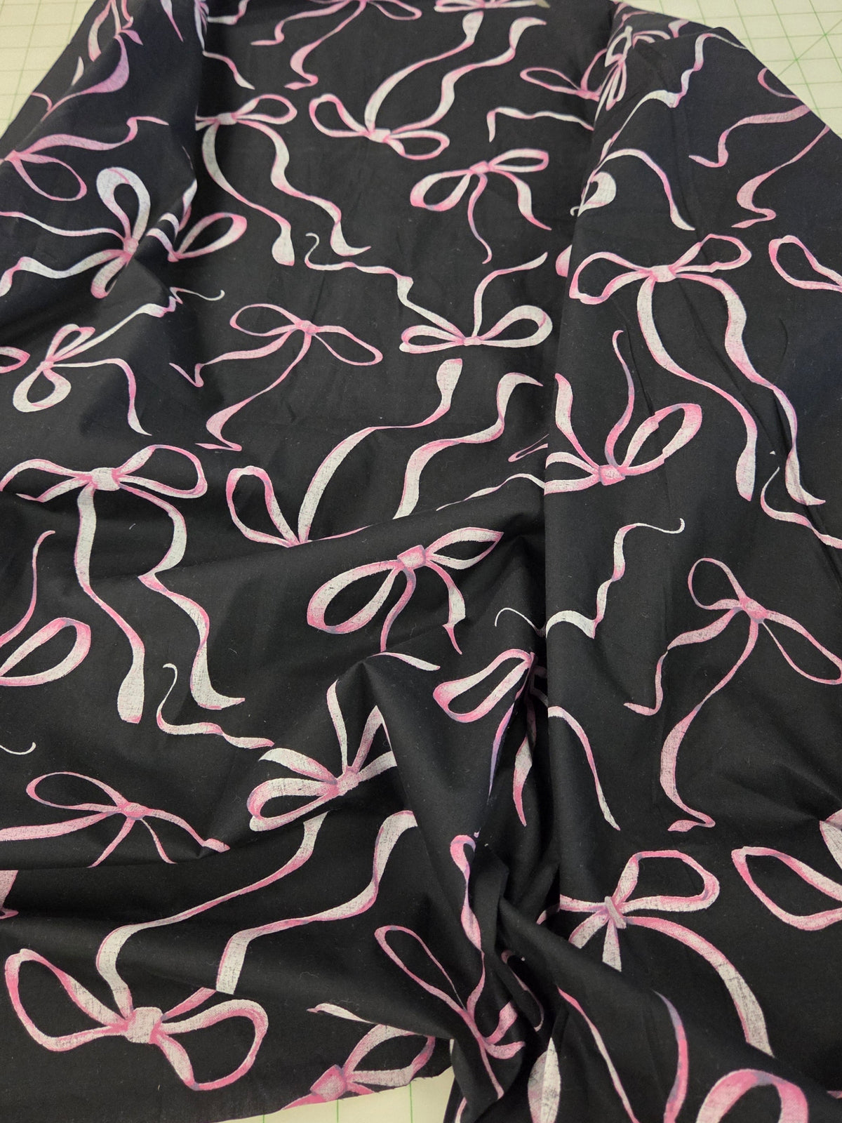 Black fabric with a pink and white bow pattern, laid out on a green cutting mat.LA FInch Fbarics. bow print ,cottage core