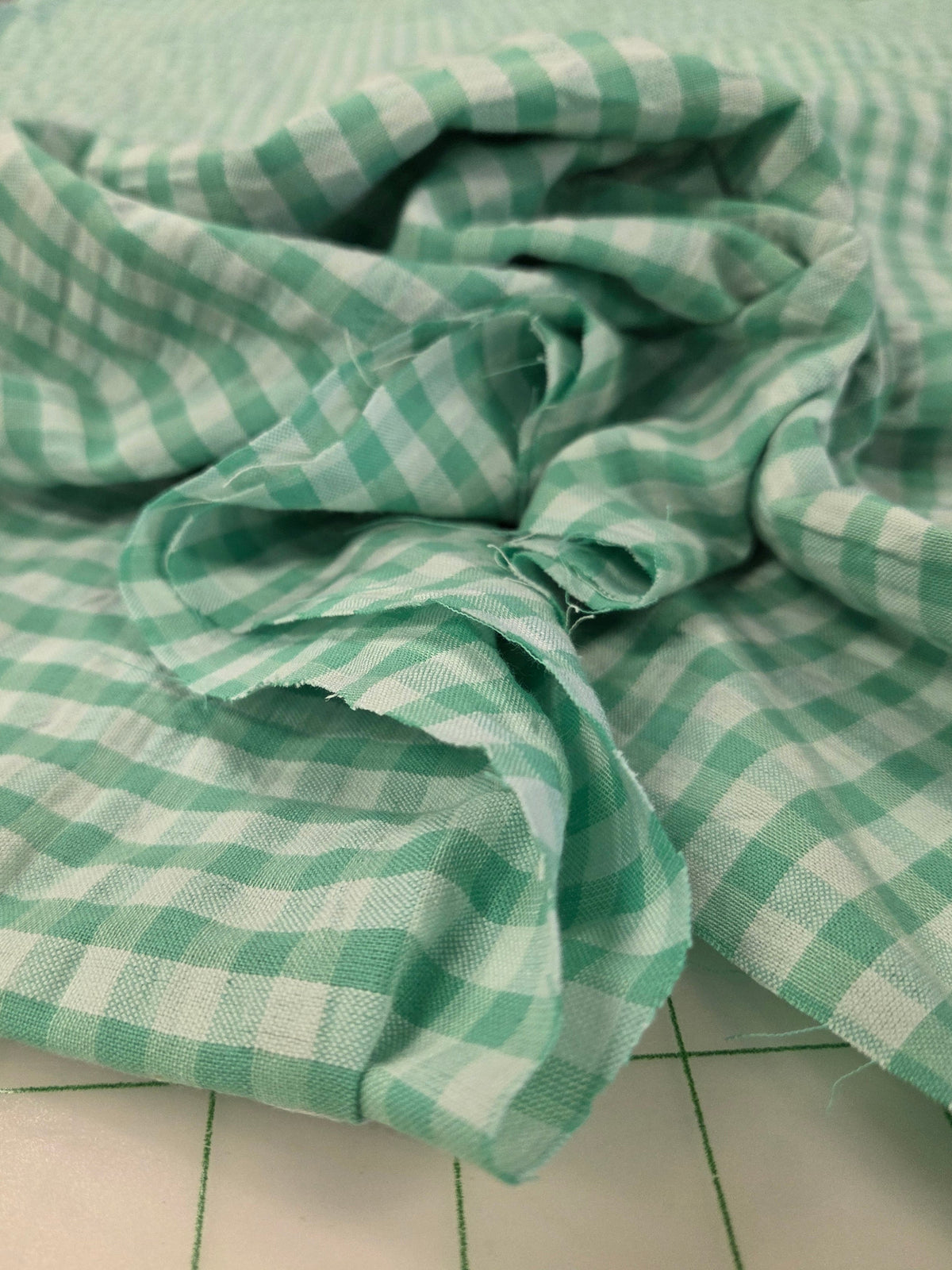 Green and white checkered fabric draped on a cutting mat, showing a soft woven texture with a gingham pattern.