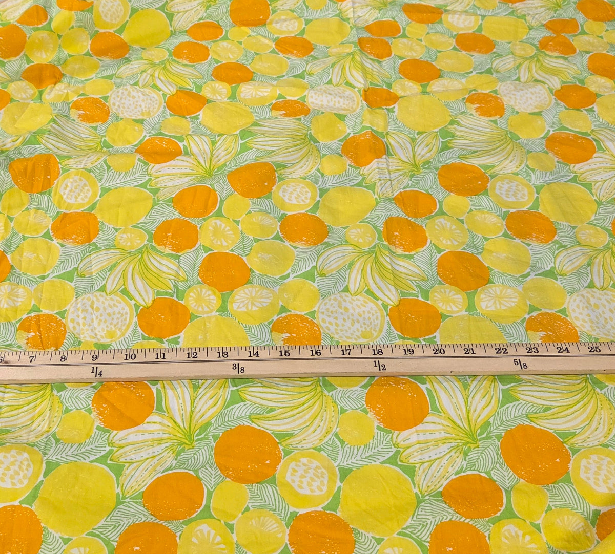 A piece of fabric with a vibrant yellow, orange, and green citrus fruit print is laid out on a white cutting mat with a wooden ruler across it, with a black pair of scissors on top. The background shows a sewing room environment with other fabrics and racks.