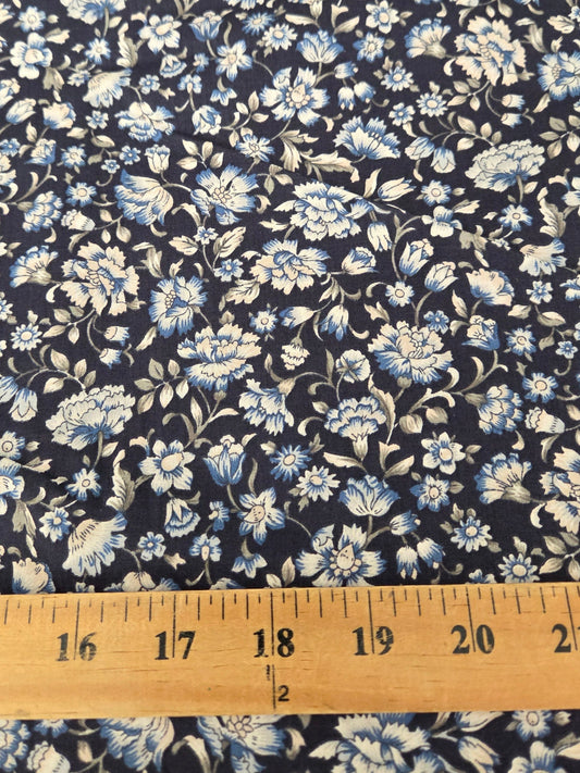 A close-up of a floral fabric with a navy blue background and light blue and white flowers, with a wooden ruler at the bottom showing the scale of the pattern.
