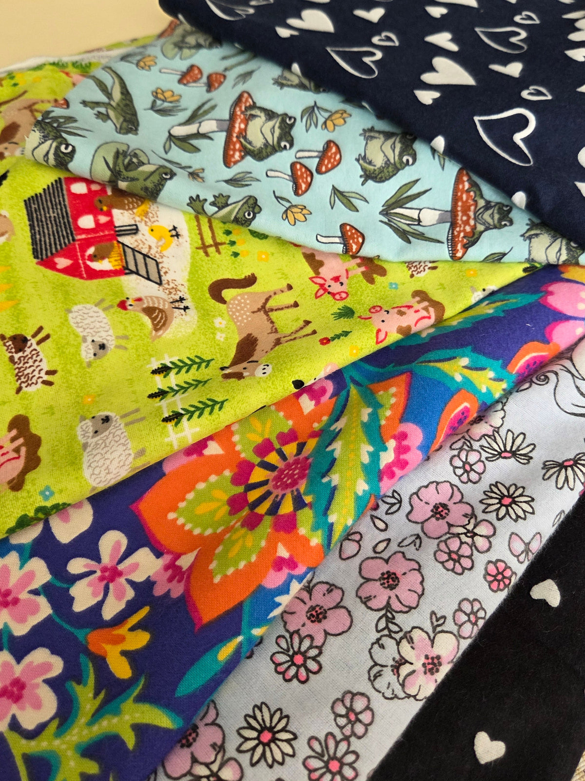 A close-up of several folded patterned fabric pieces layered on top of each other, showing different prints including white hearts on navy, woodland animals on light blue, colorful flowers, and pink floral motifs on black and white fabrics.