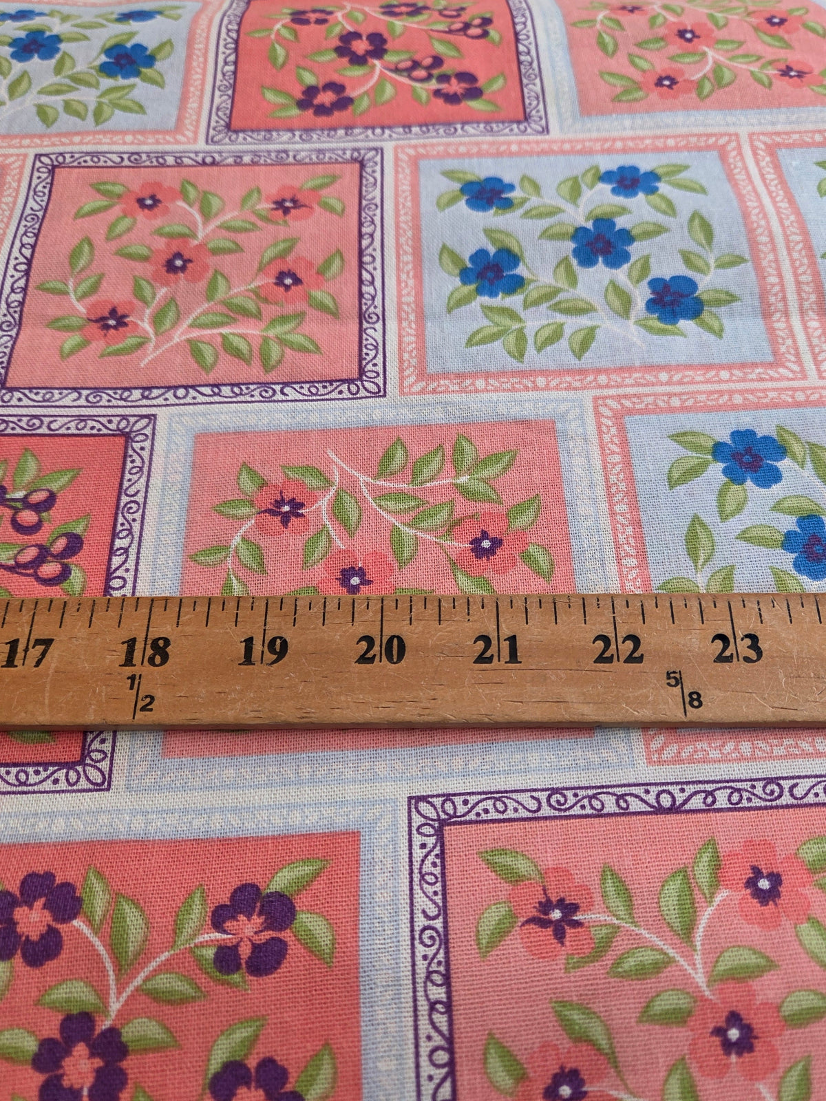 A close-up of a floral patchwork fabric with pink, light blue, and purple squares, featuring green leaves and blue and purple flowers, with a wooden ruler showing 14 inches across the bottom to indicate scale.