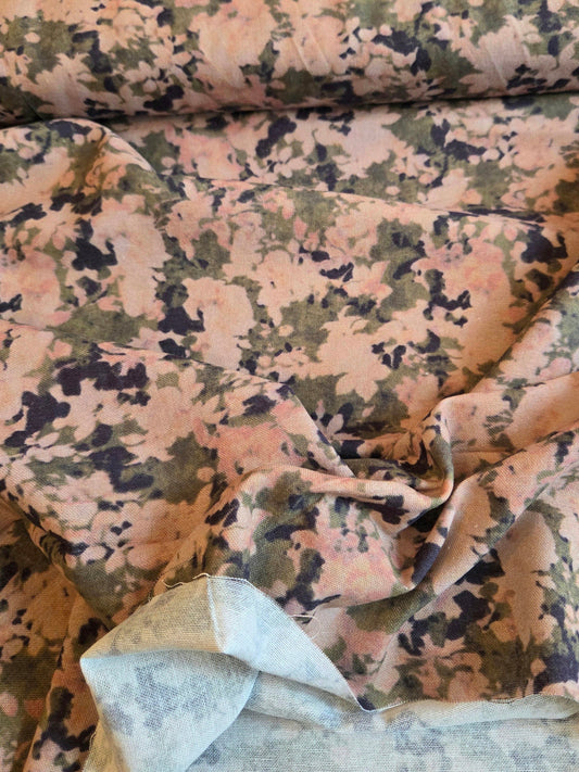 A close-up of patterned fabric with a mottled floral or camouflage-like print in pink, beige, olive green, and dark navy, with a folded section showing the reverse side.