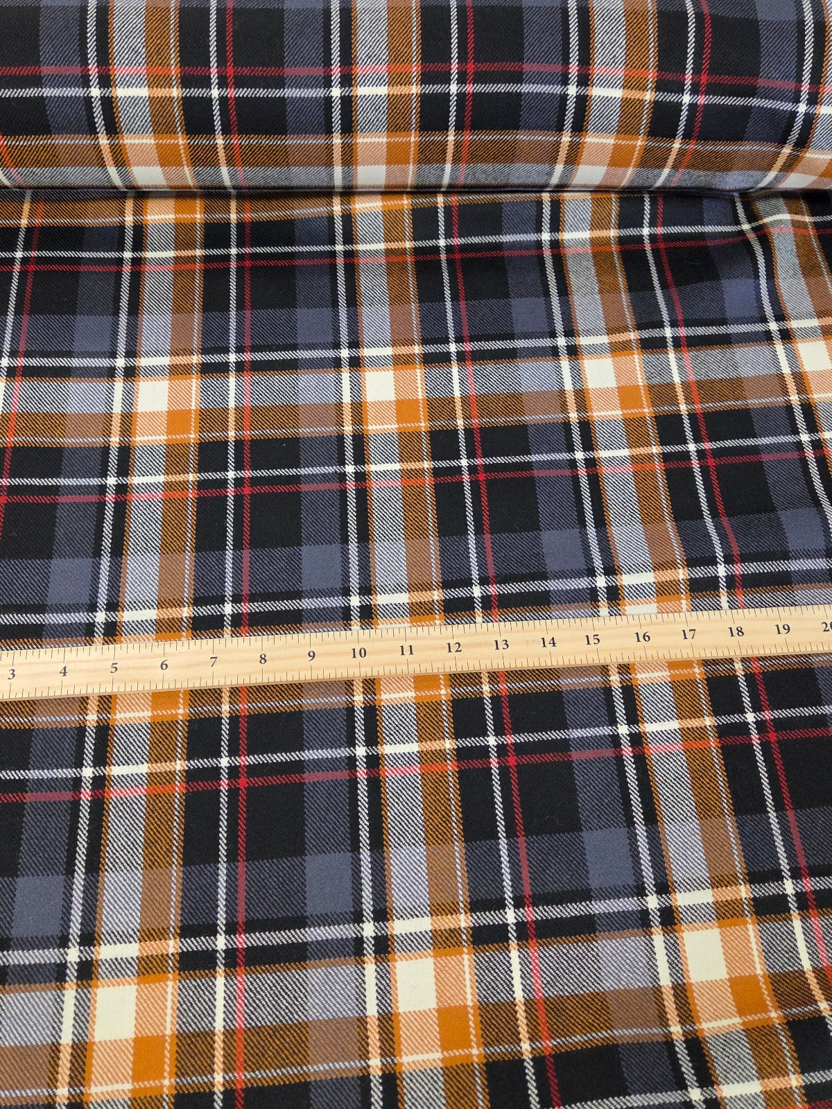 A roll of plaid fabric laid out on a cutting mat, with a wooden ruler placed across the fabric to show scale. The plaid pattern features black, navy, orange, beige, and white stripes.
