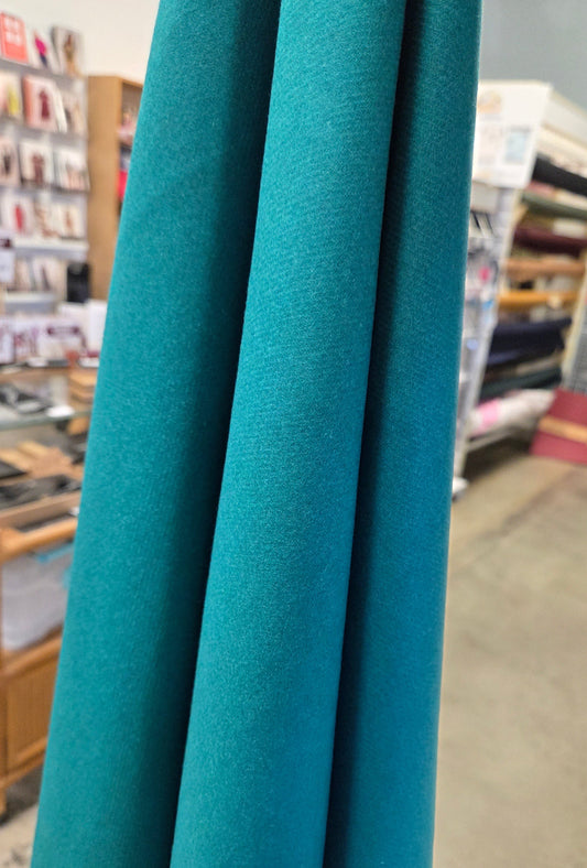 A close-up of teal blue fabric hanging vertically in folds, with a blurred store interior visible in the background. wool italian fabric coating