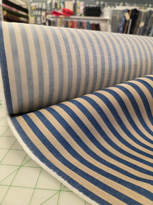 A close-up of striped fabric rolled over itself, showing alternating navy blue and warm beige vertical stripes. The fabric is photographed on a green cutting mat inside what appears to be a fabric or craft store, with shelves and hanging items blurred in the background. Cone Mills Denim fabric by the yard in Southern California. Denim Apparel Fabric Shop . LA Finch Fabrics