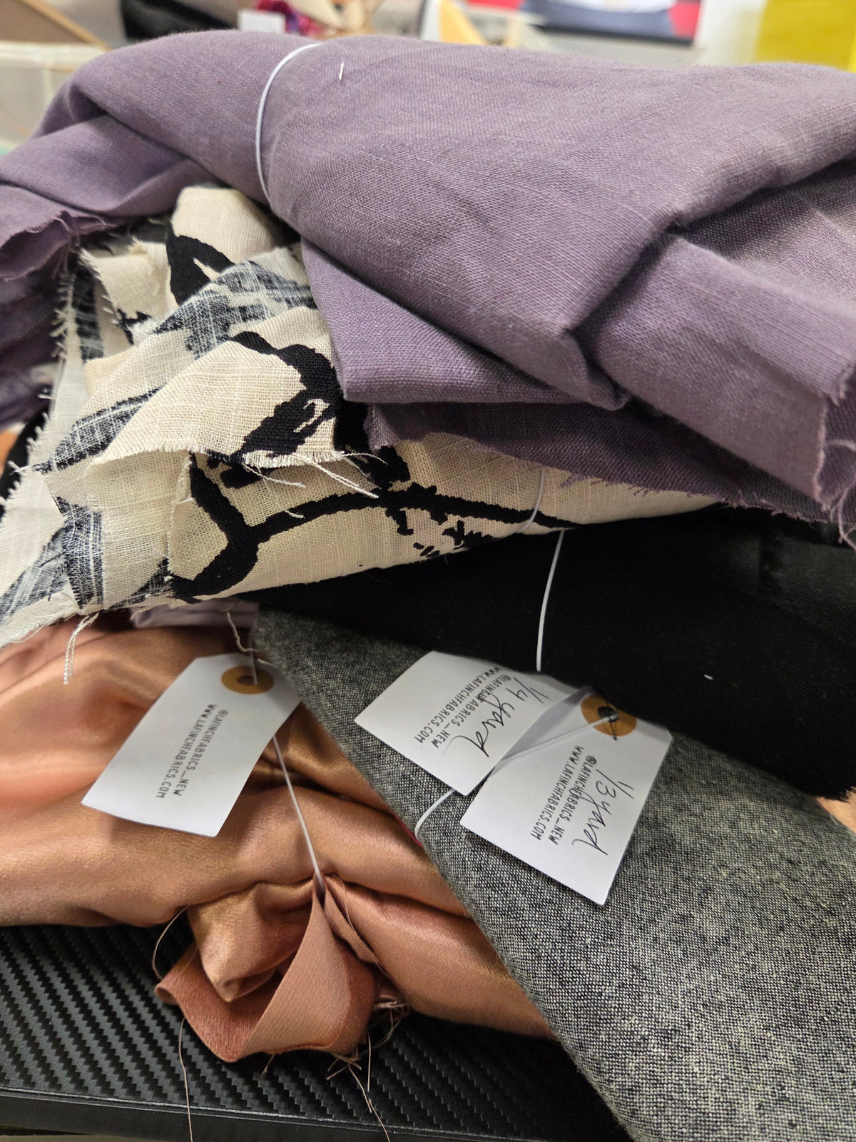 A close-up of multiple folded fabric swatches bundled together, including a lavender linen-like piece, a black fabric section, a gray wool-textured piece, a beige-and-black patterned textile, and a blush pink satin-like fabric. White tags are attached, and a measuring ruler is visible along the right edge.