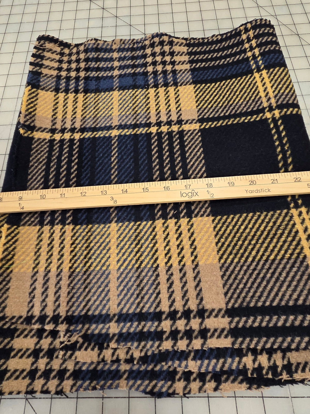 A piece of plaid fabric with navy blue, beige, and black stripes, shown on a gridded cutting mat with a wooden ruler for scale.
