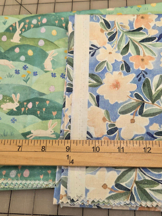 Two folded fabric bundles with floral and bunny prints laid on a gridded cutting mat, with a wooden ruler standing vertically to show measurements.