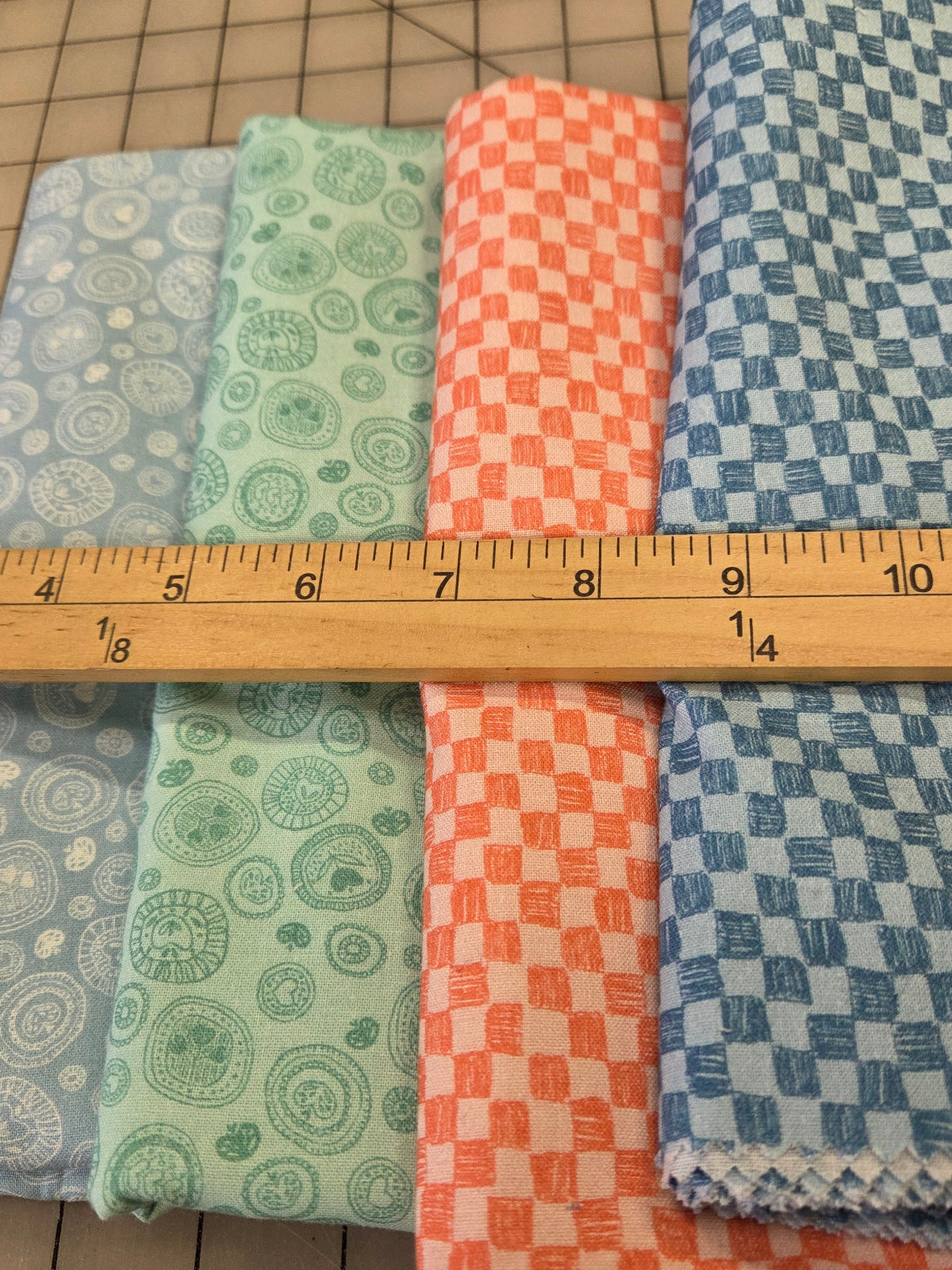 Four folded fabrics in light blue, mint green, coral orange, and medium blue with various geometric and circular patterns, laid out on a cutting mat with a wooden ruler across them.