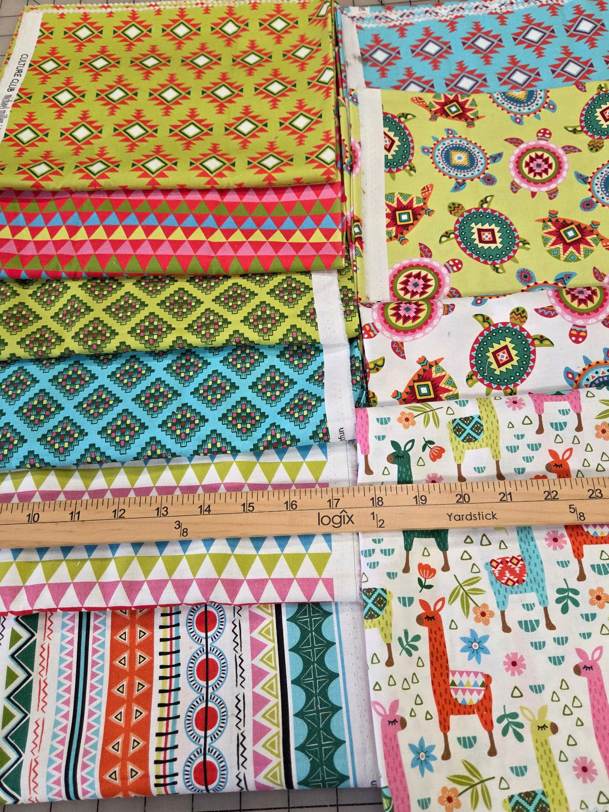 A collection of colorful patterned fabric squares arranged on a gridded cutting mat, with a wooden ruler standing vertically to show scale. The fabrics feature various geometric, floral, and tribal patterns in bright colors like yellow, blue, pink, green, and red.