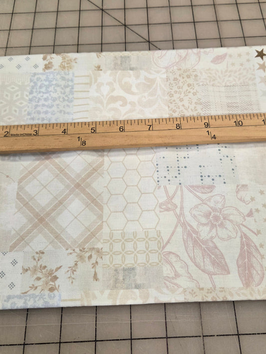 A square piece of cotton fabric with a patchwork quilt-style floral pattern in soft beige, pink, and blue tones, shown on a gridded cutting mat with a wooden ruler across the top edge for scale.