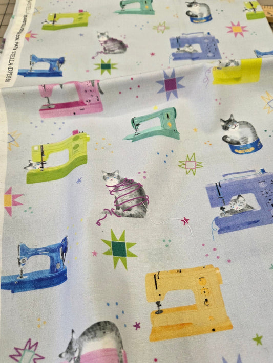 A folded piece of white fabric with a colorful print of sewing machines, cats, and starbursts, displayed on a cutting mat.