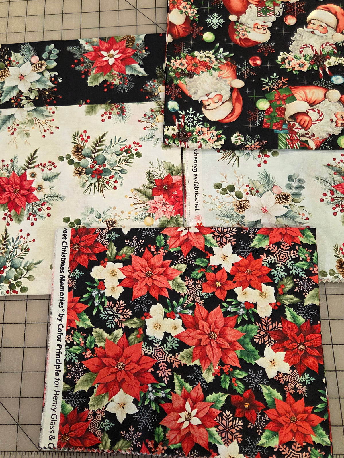 Several fabric swatches with Christmas holiday prints are arranged on a gridded cutting mat. The designs include red poinsettias, Santa Claus, snowflakes, and green foliage on black and white backgrounds, with a fabric ruler visible along the bottom edge.