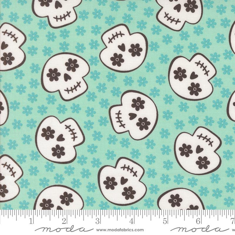 Pattern of white skulls with black details on a teal background with small flowers, ruler for scale at the bottom.