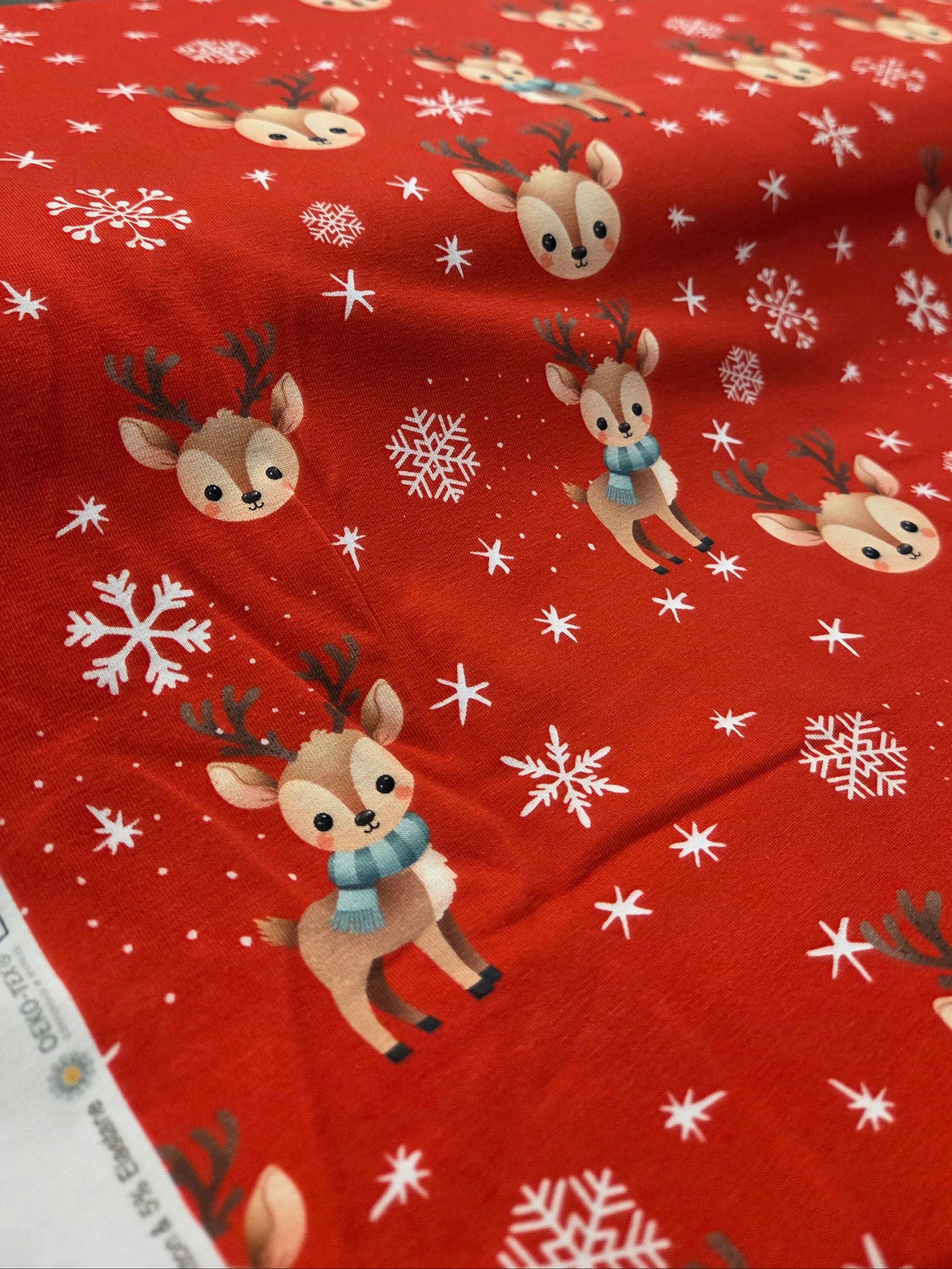 Red fabric with reindeer and snowflake pattern, possibly in a store setting. 19-1499 Holiday Fabric knit. cotton spandex Stof fabrics