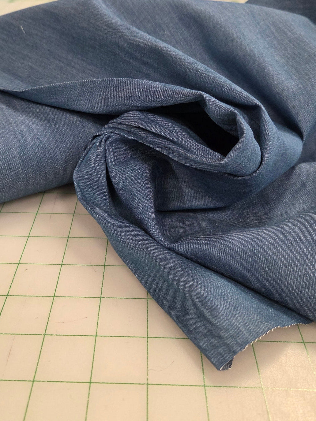 Blue fabric on a cutting mat with a store in the background. chambray light blue for sewing and making garments. sold by the yard. shirting weight cotton