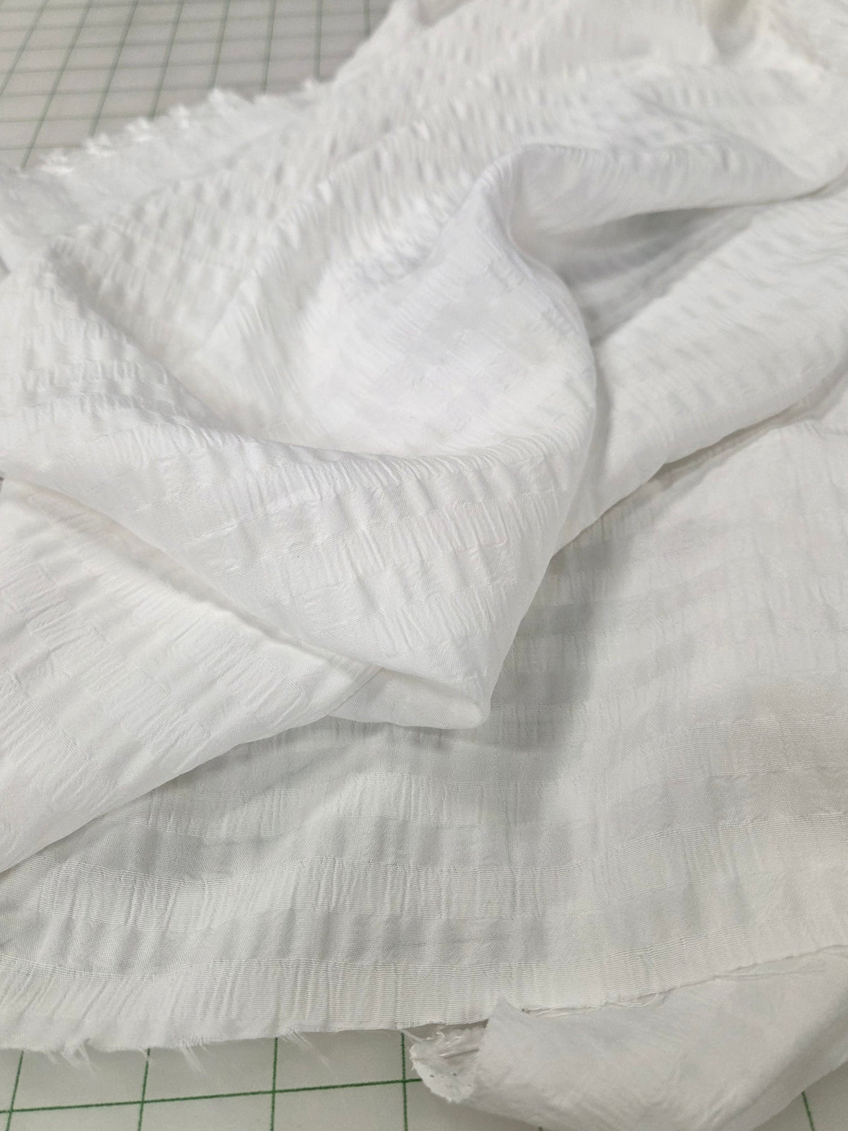 White textured fabric with a crinkled gauze-like pattern, draped and folded on a green-lined cutting mat.