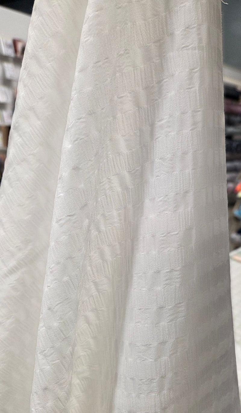 A close-up of a white textured fabric with a subtle woven pattern, hanging in folds inside what appears to be a sewing or fabric store.