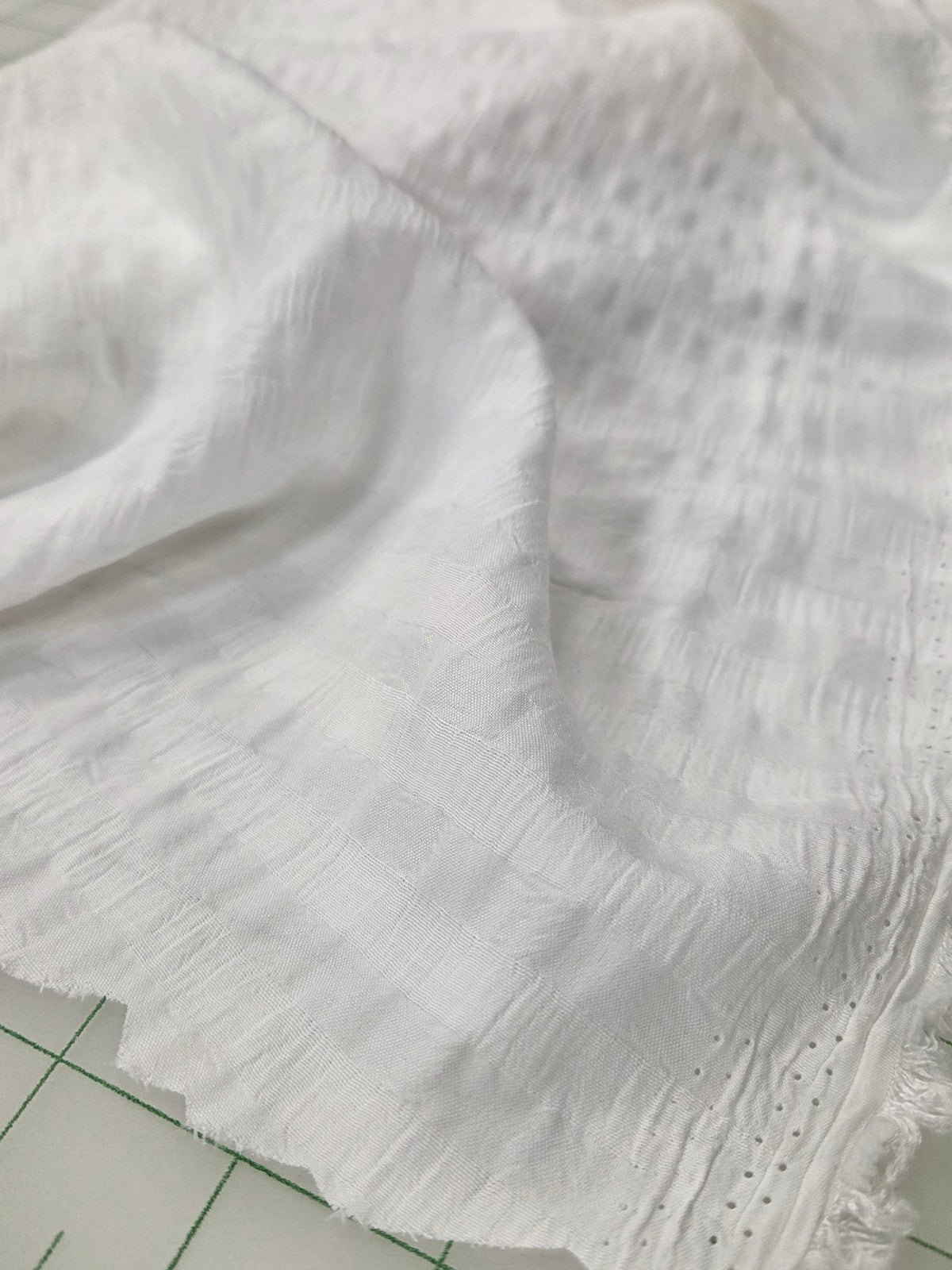 White textured fabric with a frayed hem and small eyelet details, draped on a green grid cutting mat.
