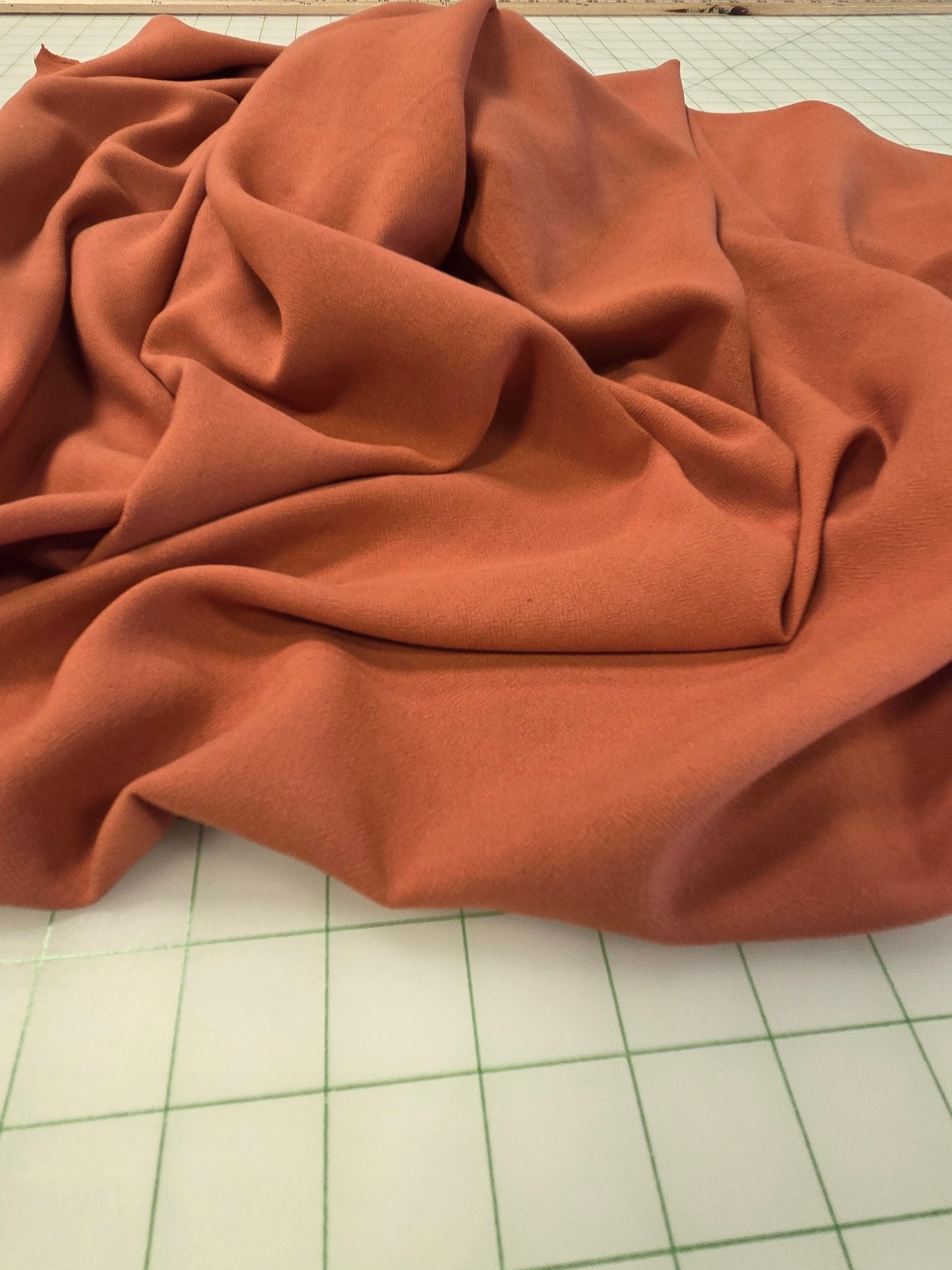 A draped piece of rust-colored fabric spread out on a white grid cutting mat, with shelves of other fabrics visible in the background.
