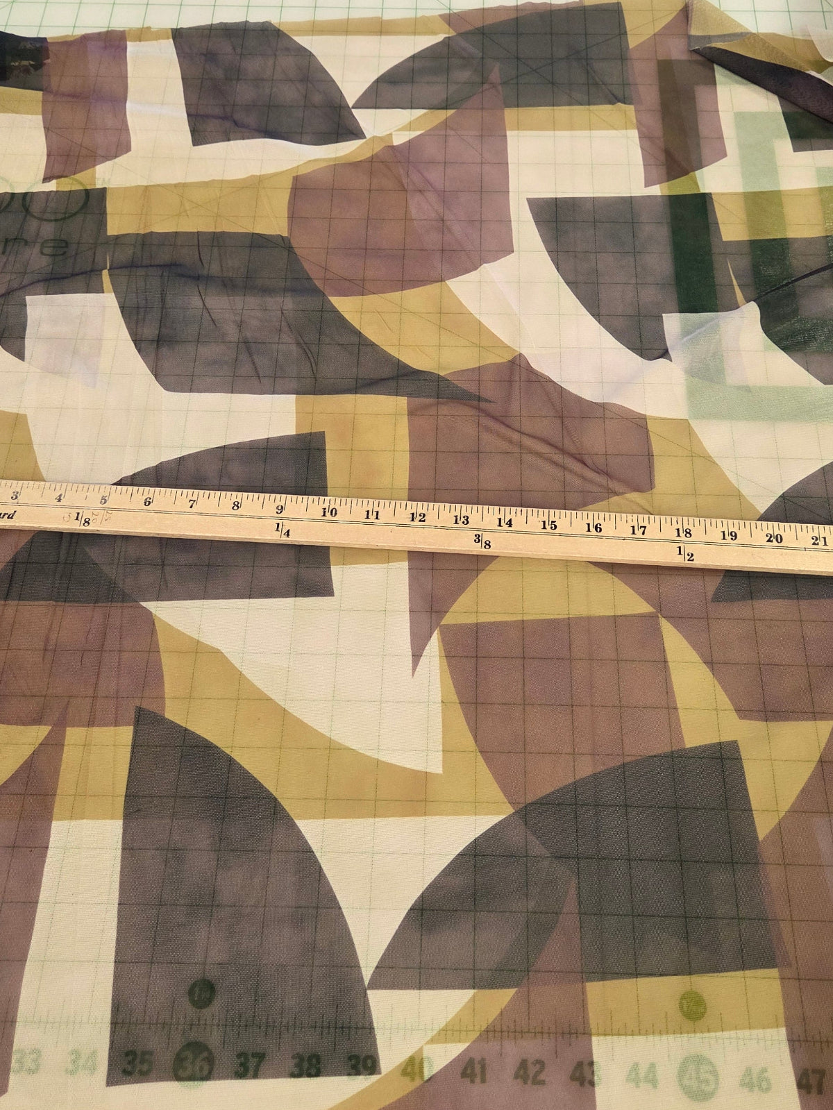 A large fabric yardage with an abstract geometric pattern in brown, beige, olive green, and black, laid flat on a white cutting mat with a wooden ruler placed vertically for scale.