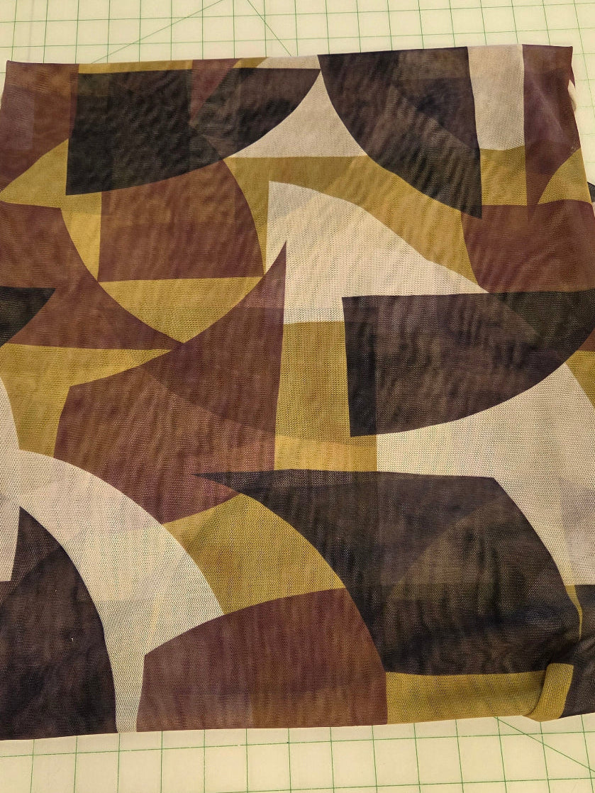 A square piece of patterned fabric with an abstract geometric design in brown, beige, white, and dark gray, laid flat on a green grid cutting mat.