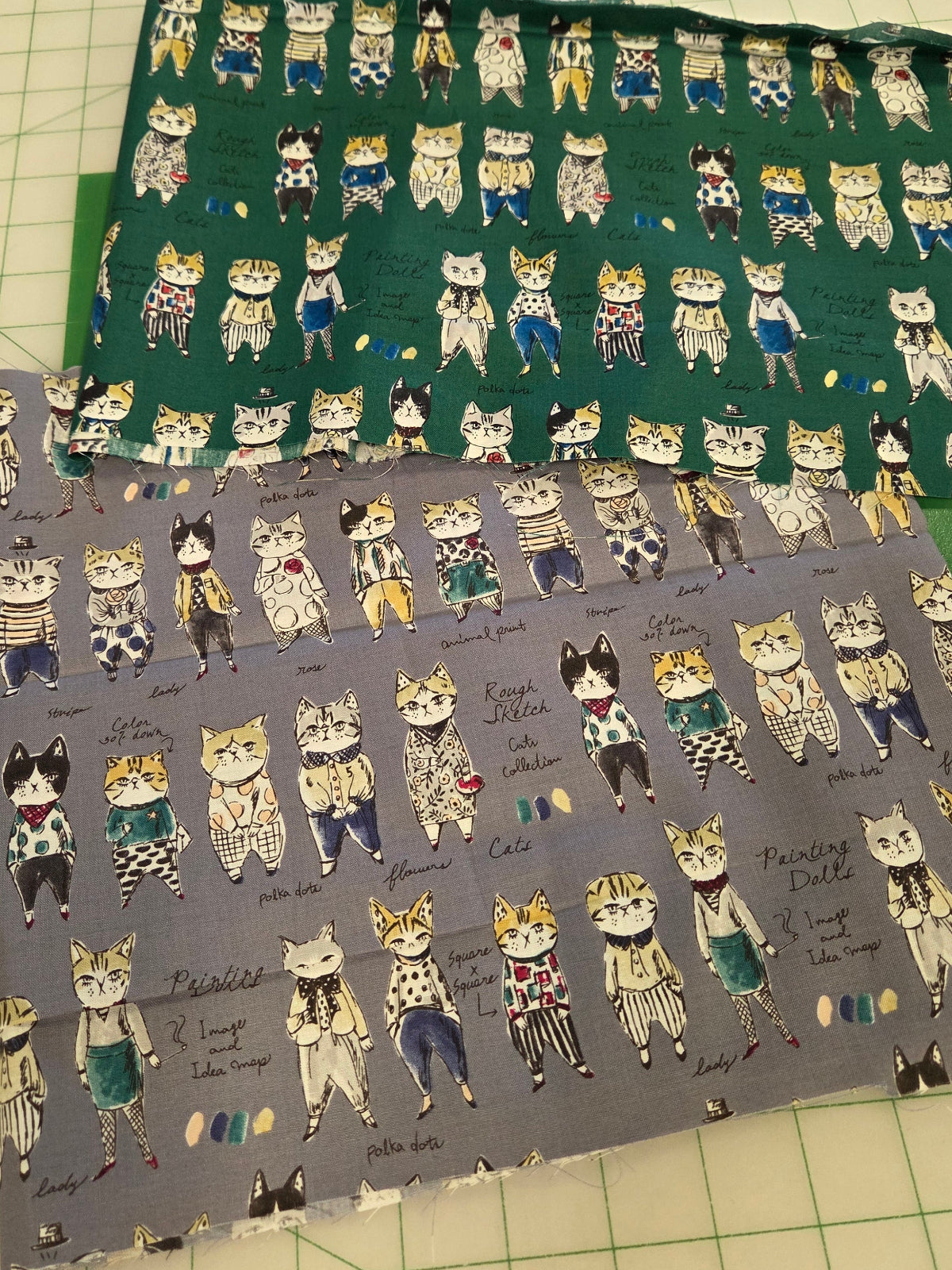 Two fabric pieces with cartoon cat illustrations, one green and one gray, laid flat on a green cutting mat.