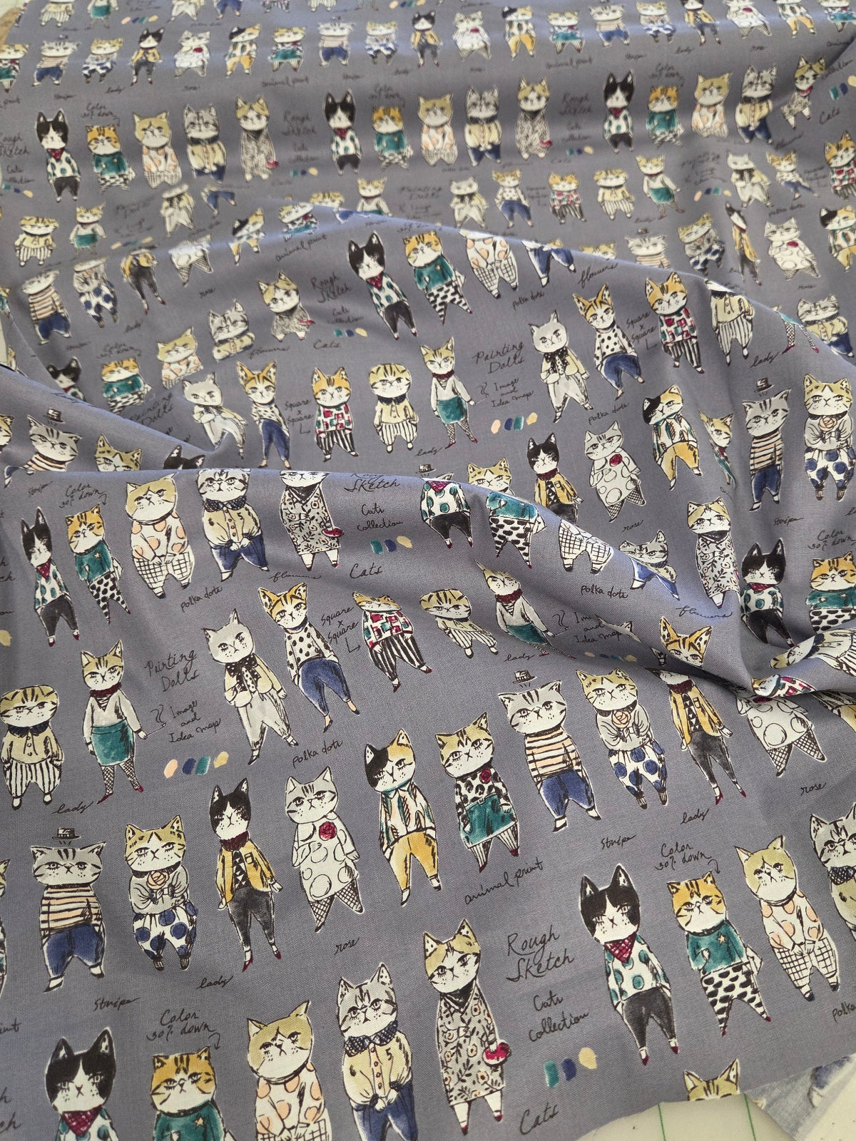 A piece of gray fabric printed with a repeating pattern of cute illustrated cats in various poses and colors, laid out on a green grid cutting mat.