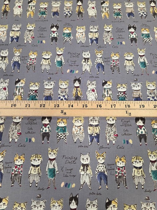 A close-up of patterned fabric with rows of illustrated cats in different outfits on a gray background, with a wooden ruler laid horizontally to show scale.