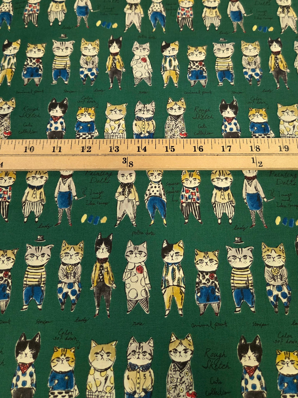 Green fabric with a repeating pattern of illustrated cats in various outfits, shown with a wooden ruler across the middle for scale.