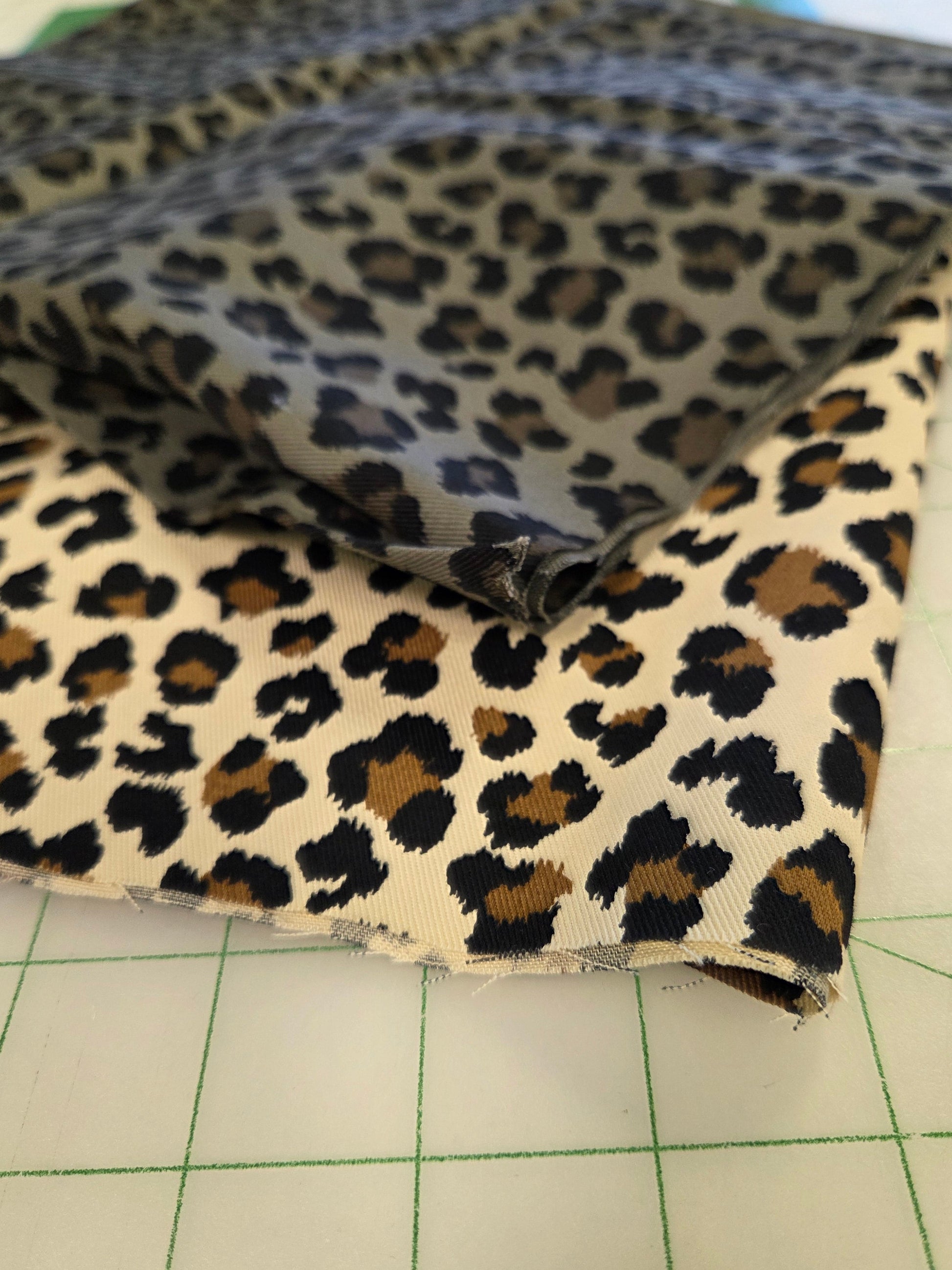 Leopard print fabric draped over a white gridded cutting mat, showing a black, brown, and beige animal spot pattern.