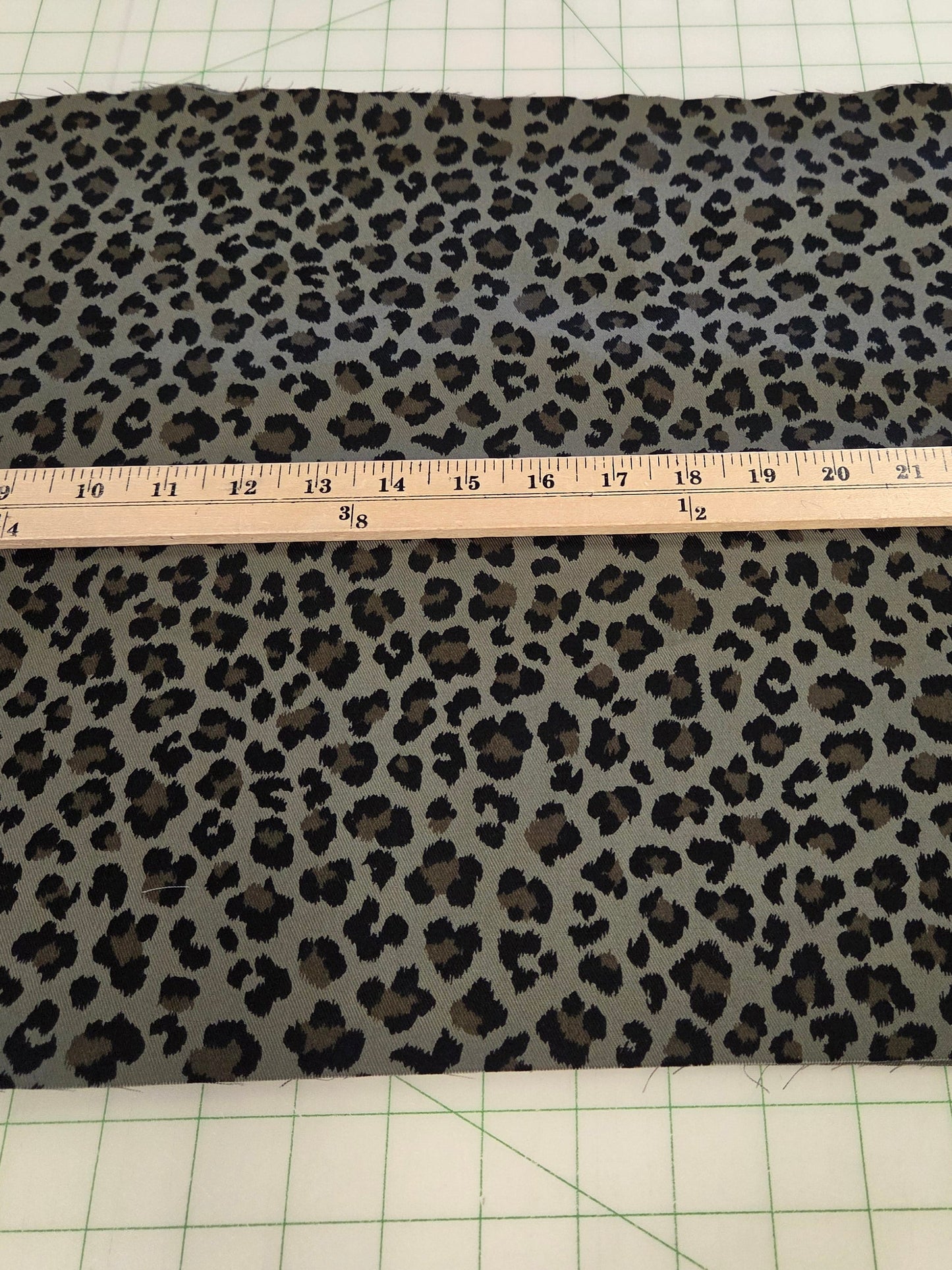A rectangular piece of leopard print fabric laid flat on a green grid cutting mat, with a wooden ruler placed across the center to show the material width.
