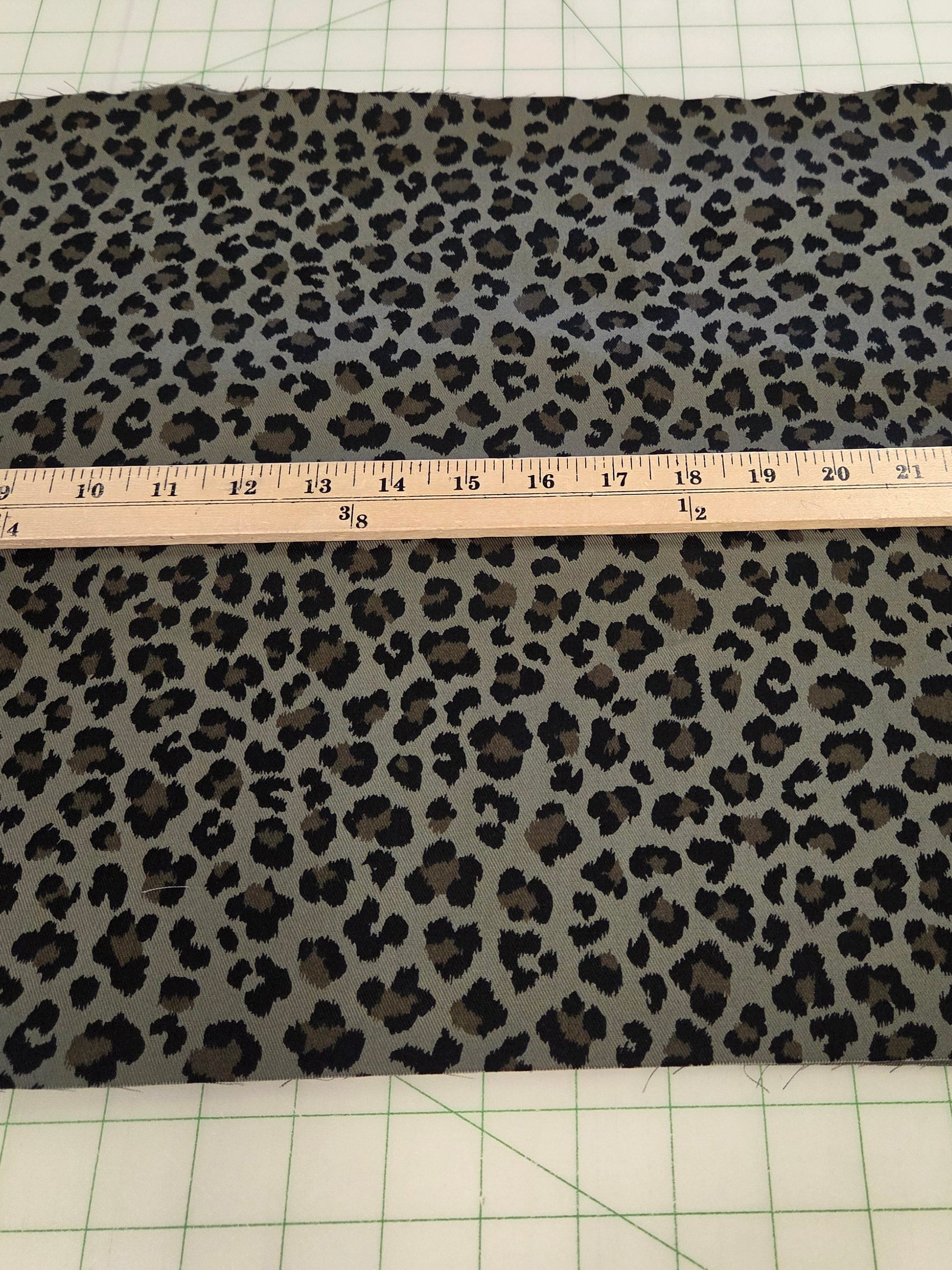 A rectangular piece of leopard print fabric laid flat on a green grid cutting mat, with a wooden ruler placed across the center to show the material width.